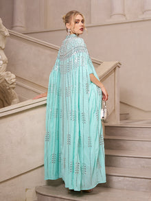 A woman in the Social Queen Crystal-Embellished Cape Dress stands on a staircase, looking over her shoulder and holding a small silver purse.