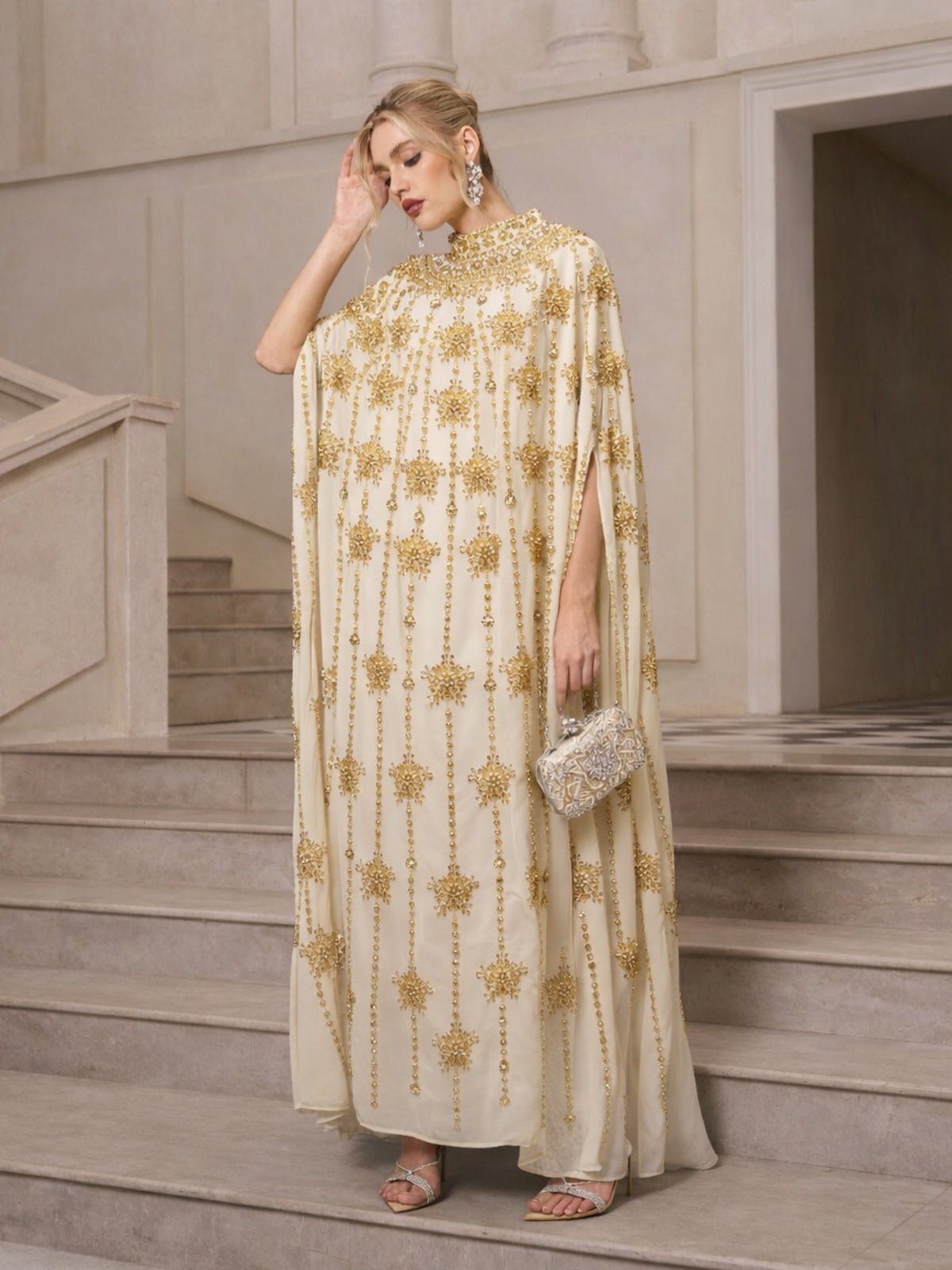 A woman stands on stairs in the Social Queen Crystal-Embellished Cape Dress, paired with statement earrings, silver heels, and a silver clutch.