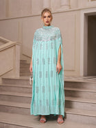 A woman in a turquoise, floor-length Crystal-Embellished Cape Dress by Social Queen stands on stone stairs, holding a small silver handbag and wearing silver heels.