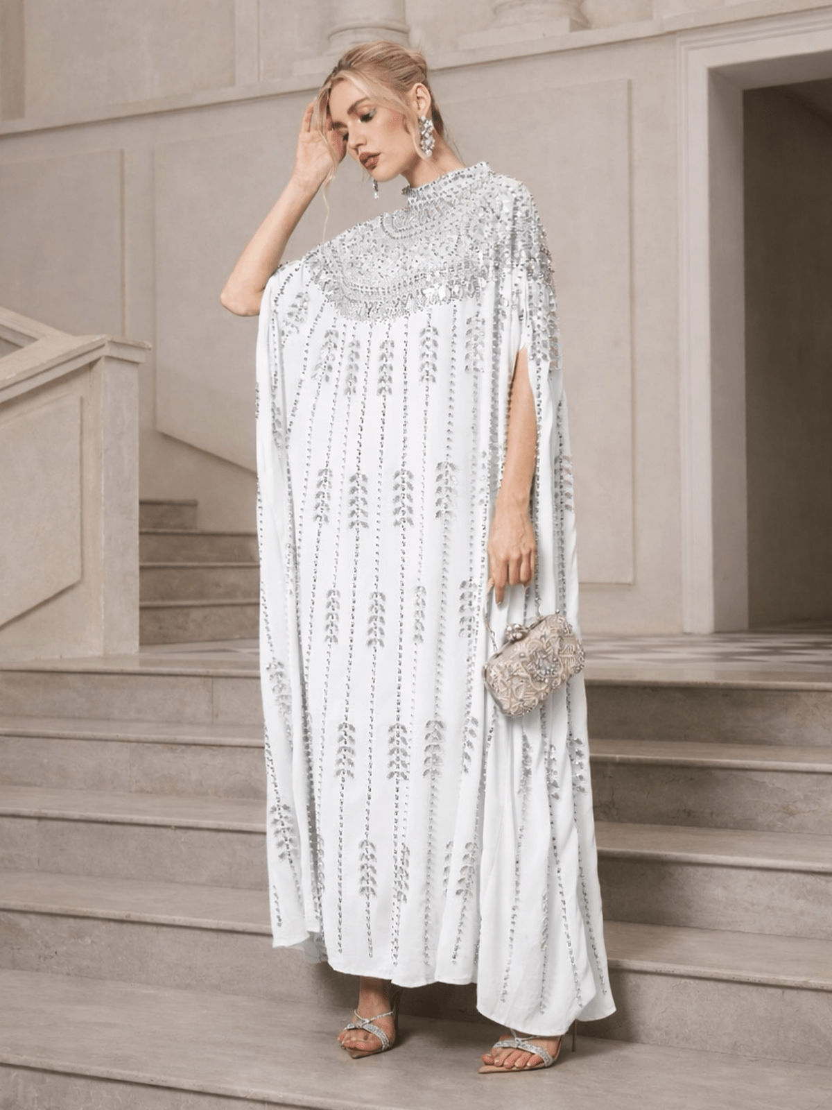 A woman in a Social Queen Crystal-Embellished Cape Dress, silver sandals, and statement earrings stands on stone steps holding a silver clutch.