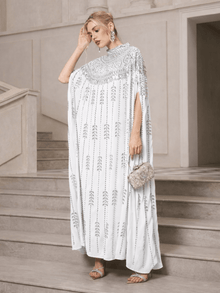 A woman in a Social Queen Crystal-Embellished Cape Dress, silver sandals, and statement earrings stands on stone steps holding a silver clutch.