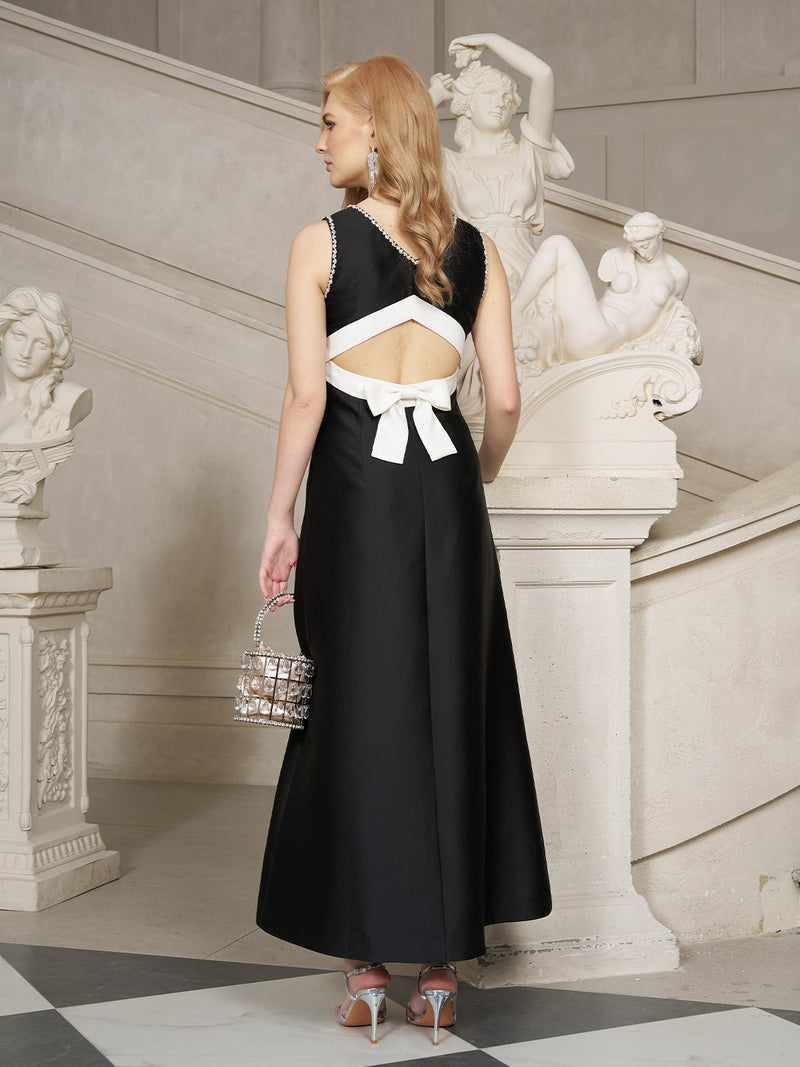 A woman stands on a marble staircase holding a metallic handbag, wearing Social Queen’s Crystal-Embellished Dress with Bow Open Back. Marble statues can be seen in the background.