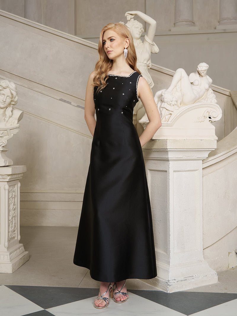 A woman wears the Social Queen Crystal-Embellished Dress with Bow Open Back, standing before classical sculptures and a staircase, gazing to the side with her hands behind her back.