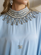 A woman wears the Social Queen Crystal-Embellished Mock-Neck Caftan Dress, featuring light blue fabric, an ornate jeweled collar, and a decorative chest brooch.