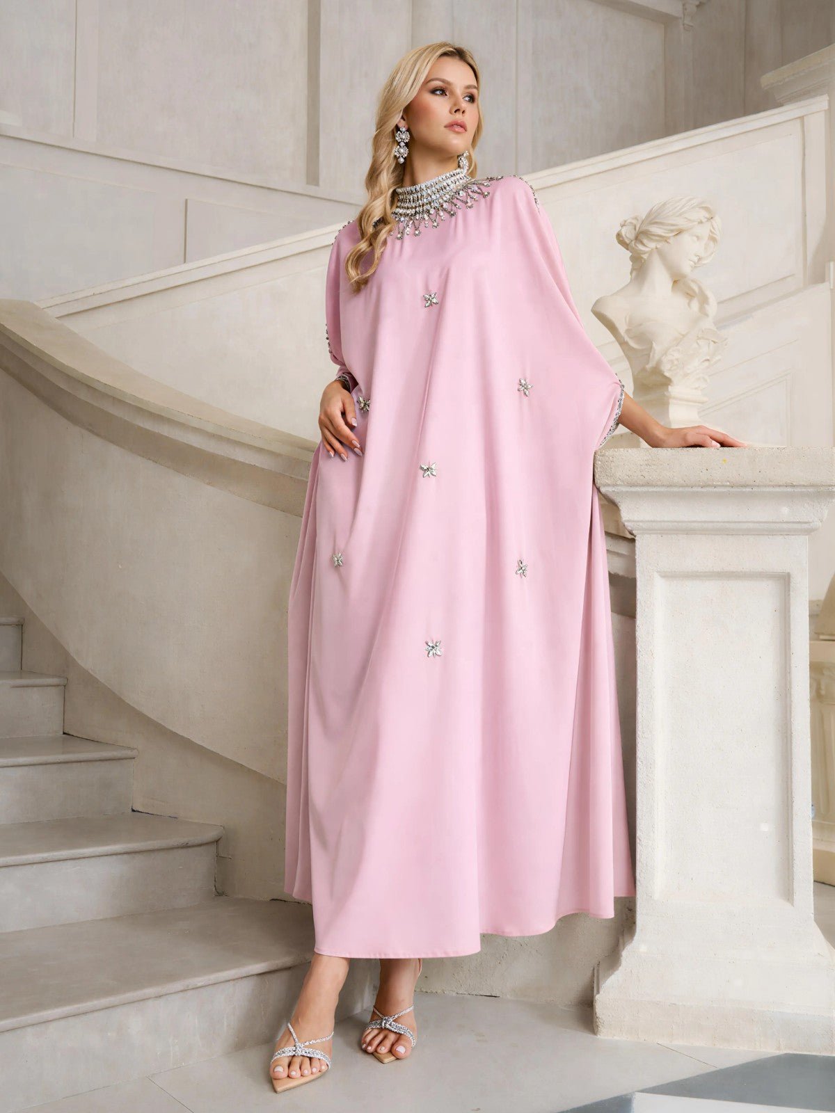 A woman stands on a staircase next to a white statue, wearing the Social Queen Crystal-Embellished Mock-Neck Caftan Dress in light pink, paired with silver sandals and jewelry.