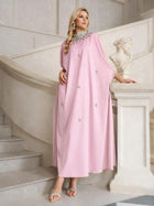 A woman stands on a staircase next to a white statue, wearing the Social Queen Crystal-Embellished Mock-Neck Caftan Dress in light pink, paired with silver sandals and jewelry.