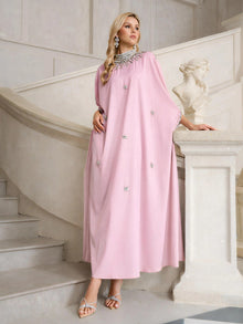 A woman stands on a staircase next to a white statue, wearing the Social Queen Crystal-Embellished Mock-Neck Caftan Dress in light pink, paired with silver sandals and jewelry.