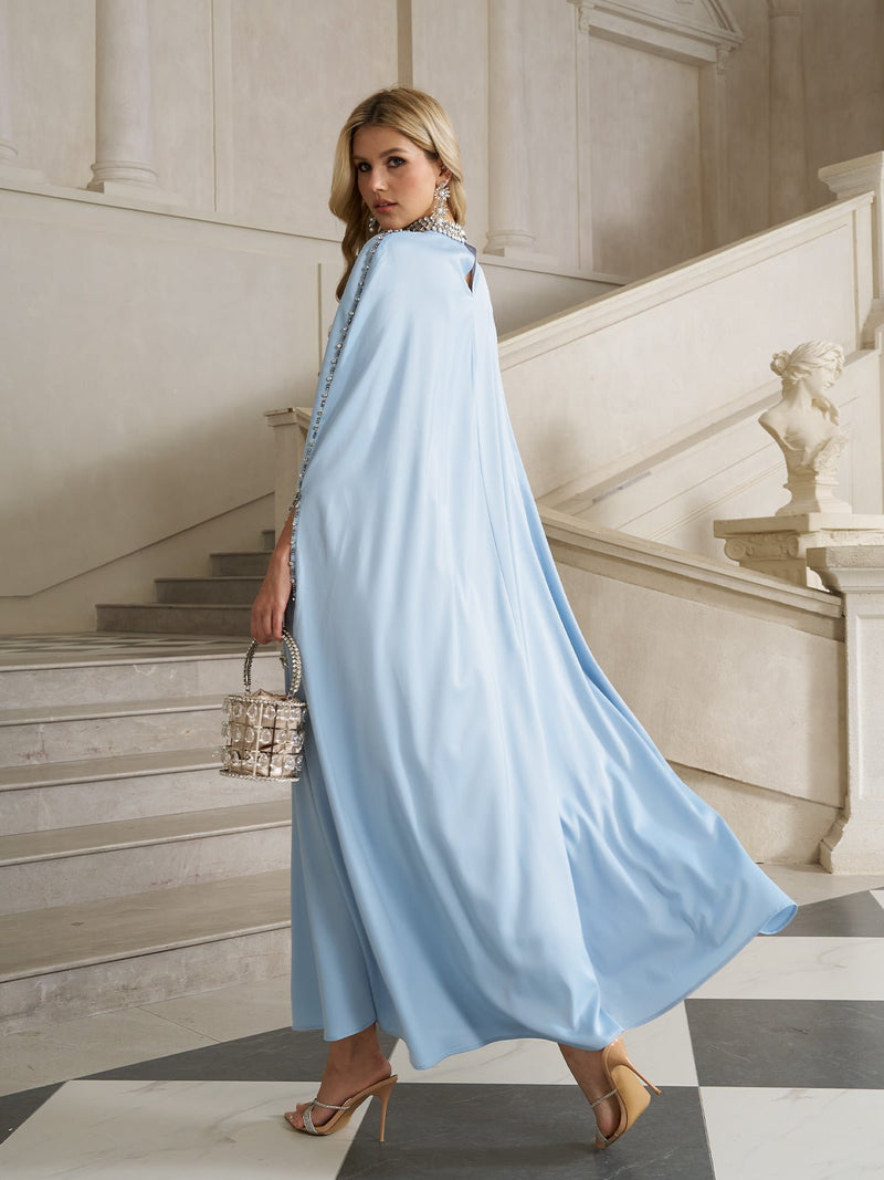 A woman in a Social Queen Crystal-Embellished Mock-Neck Caftan Dress and heels stands on marble stairs, holding a small handbag and glancing over her shoulder in an elegant, classic interior.