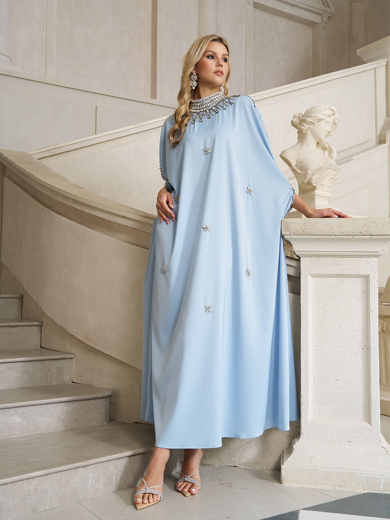 A woman in the Crystal-Embellished Mock-Neck Caftan Dress by Social Queen stands on a marble staircase beside a white bust, set in an elegant interior.