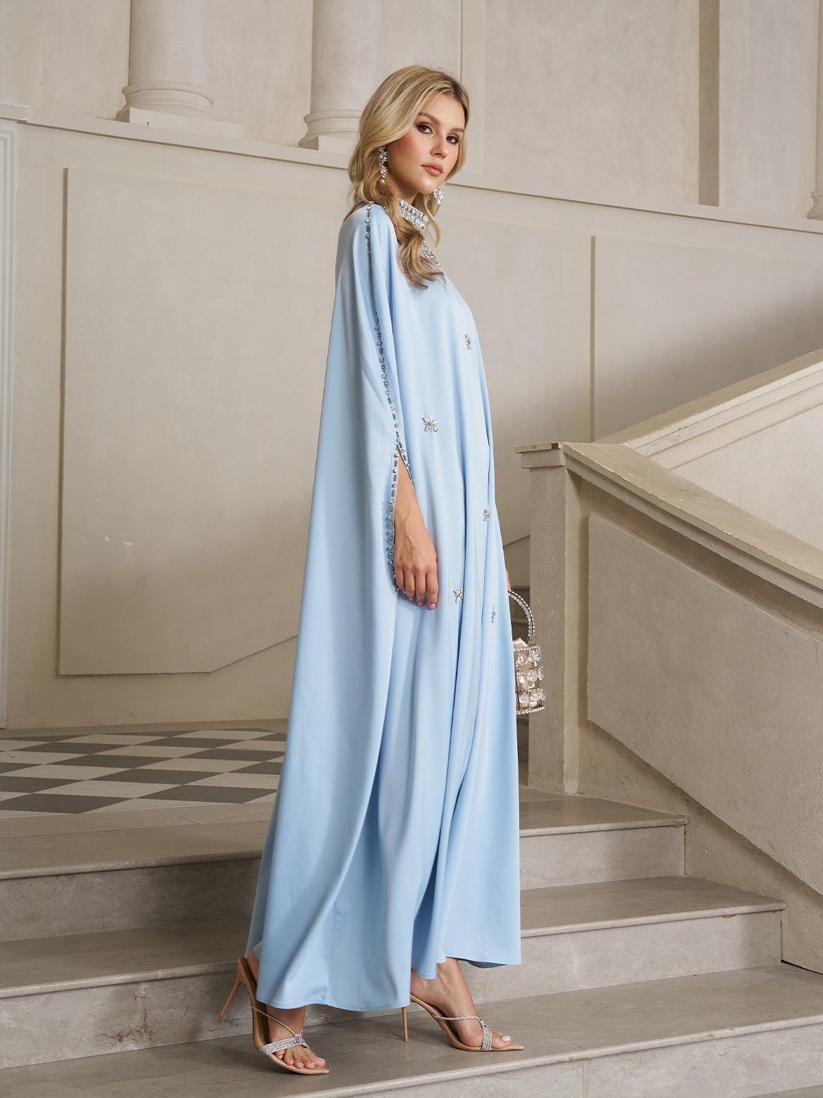 A woman stands on stairs in an elegant, neutral-toned interior, wearing the Social Queen Crystal-Embellished Mock-Neck Caftan Dress with high-heeled sandals and carrying a transparent handbag.