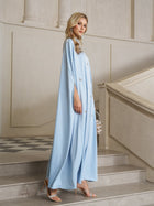 A woman stands on stairs in an elegant, neutral-toned interior, wearing the Social Queen Crystal-Embellished Mock-Neck Caftan Dress with high-heeled sandals and carrying a transparent handbag.