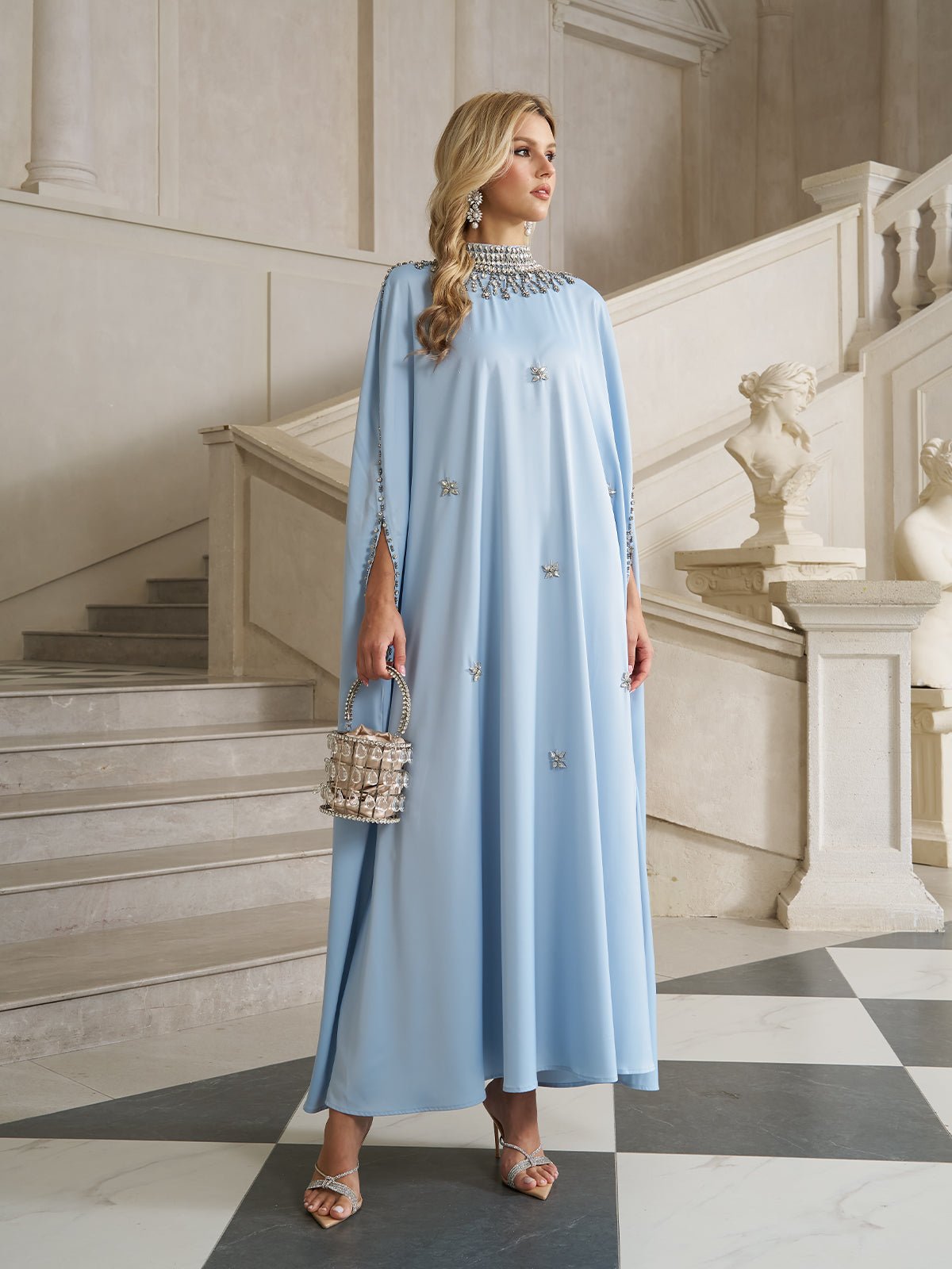 A woman in a Crystal-Embellished Mock-Neck Caftan Dress by Social Queen stands by a staircase indoors, holding a small handbag and wearing strappy heels. Marble statues and ornate architecture are in the background.