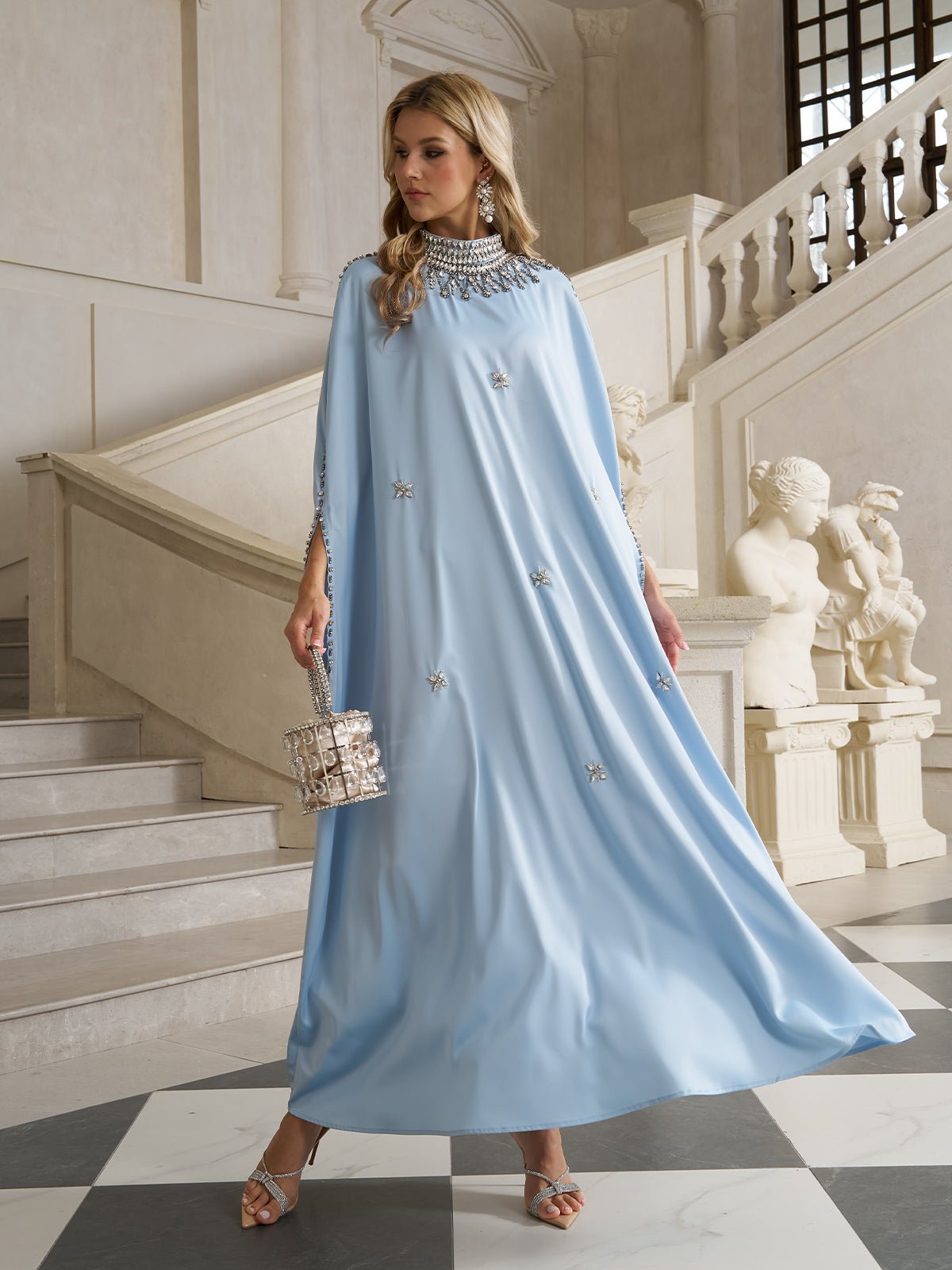 A woman in the Social Queen Crystal-Embellished Mock-Neck Caftan Dress stands on an ornate staircase, holding a transparent box-shaped handbag, surrounded by marble statues and classical architecture.