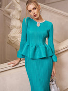 A woman in the Social Queen Crystal-Embellished Pleated Peplum Set stands by a staircase holding a silver handbag, with a white statue behind her—a chic wedding guest look.