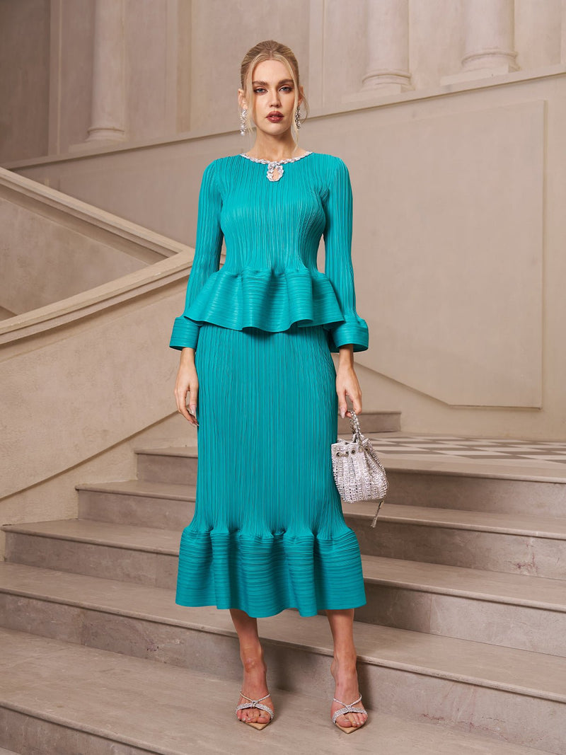 A woman wears the Social Queen Crystal-Embellished Pleated Peplum Set—a teal, long-sleeved top and matching fishtail skirt—paired with a silver handbag and strappy heels, creating an elegant wedding guest look.
