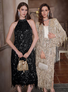 Two women stand side by side: one in Social Queen’s Crystal-Embellished Sequin Fringe Halter Dress with a gold purse, the other in a gold fringe outfit and silver clutch. Both are elegantly dressed for a formal event.