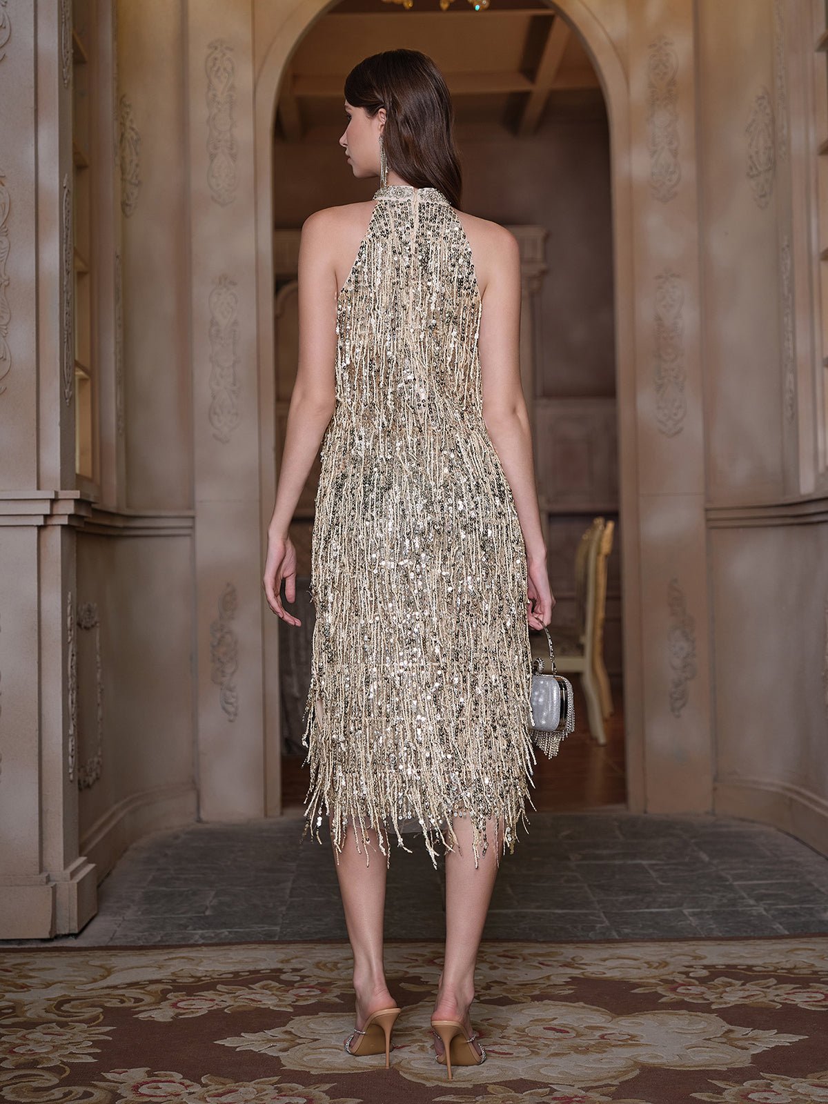 A woman stands indoors facing away, wearing the Social Queen Crystal-Embellished Sequin Fringe Halter Dress with heels and a small handbag—ideal for formal events.