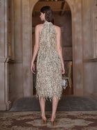 A woman stands indoors facing away, wearing the Social Queen Crystal-Embellished Sequin Fringe Halter Dress with heels and a small handbag—ideal for formal events.
