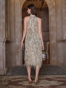 A woman stands indoors facing away, wearing the Social Queen Crystal-Embellished Sequin Fringe Halter Dress with heels and a small handbag—ideal for formal events.