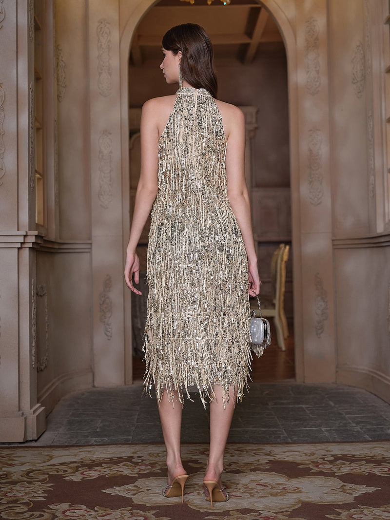 A woman stands indoors facing away, wearing the Social Queen Crystal-Embellished Sequin Fringe Halter Dress with heels and a small handbag—ideal for formal events.
