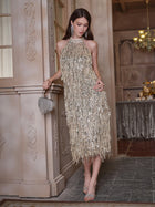 A woman stands indoors wearing the Social Queen Crystal-Embellished Sequin Fringe Halter Dress, paired with matching earrings and heels, holding a small fringed purse in an elegantly decorated room.