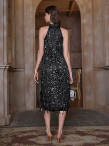 A woman stands indoors facing away, wearing the Social Queen Crystal-Embellished Sequin Fringe Halter Dress and heeled sandals, holding a small silver purse.