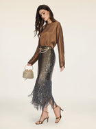 A woman stands against a plain background wearing the Social Queen Crystal-Embellished Tencel Satin Blouse, a metallic fringed skirt, gold heels, and holding a beaded purse.
