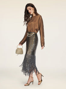 A woman stands against a plain background wearing the Social Queen Crystal-Embellished Tencel Satin Blouse, a metallic fringed skirt, gold heels, and holding a beaded purse.