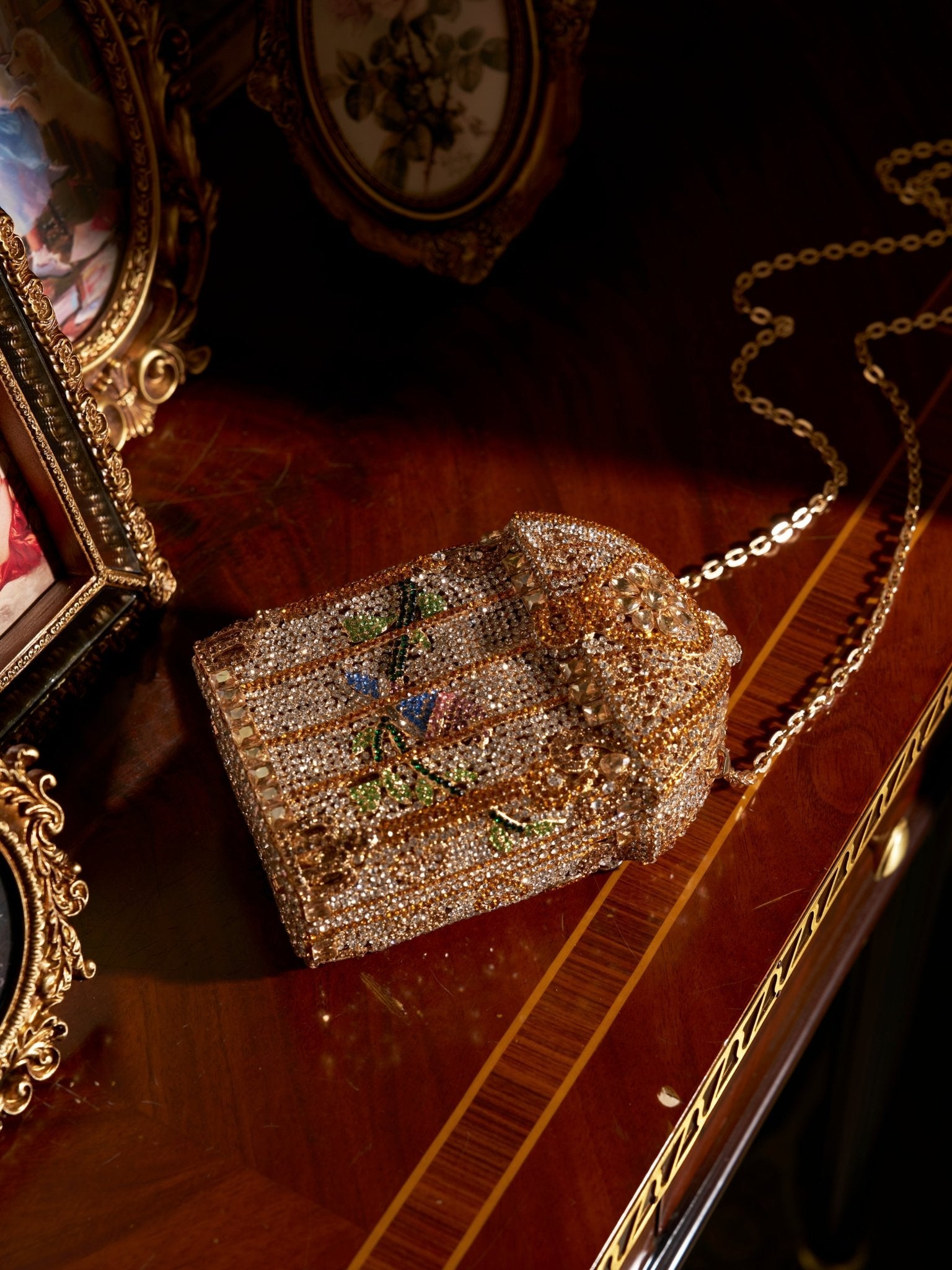 The Crystal-Embellished Treasure Box Bag by Social Queen, featuring a gold chain strap and ornate rhinestones, rests on polished wood beside framed photos, its sparkling details catching the light.