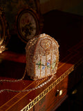 The Crystal-Embellished Treasure Box Bag by Social Queen, a jeweled vintage-inspired handbag with a chain strap, sits on a polished wooden table next to two framed floral pictures.