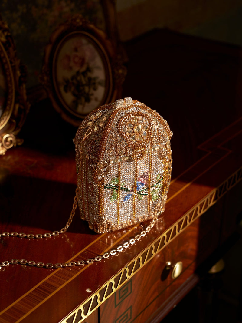 The Crystal-Embellished Treasure Box Bag by Social Queen, a jeweled vintage-inspired handbag with a chain strap, sits on a polished wooden table next to two framed floral pictures.