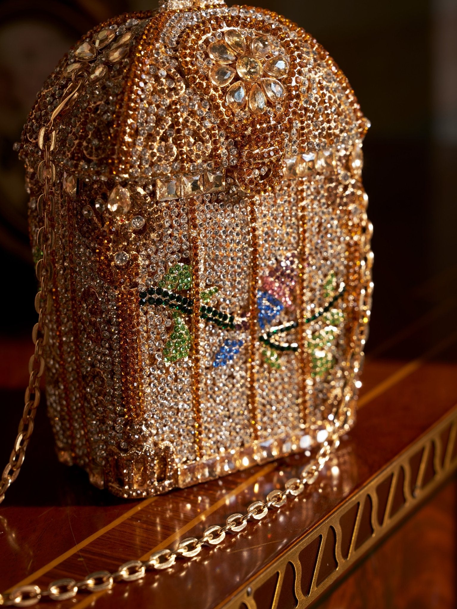 The Crystal-Embellished Treasure Box Bag by Social Queen, featuring a birdcage motif with colorful birds, rhinestones, and floral accents, rests elegantly on a wooden surface.