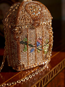 The Crystal-Embellished Treasure Box Bag by Social Queen, featuring a birdcage motif with colorful birds, rhinestones, and floral accents, rests elegantly on a wooden surface.