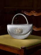 The Social Queen Crystal Floral Satin Mini Top-Handle Bag, featuring a crystal floral embellishment and satin finish, rests on the yellow seat of a carved wooden chair.