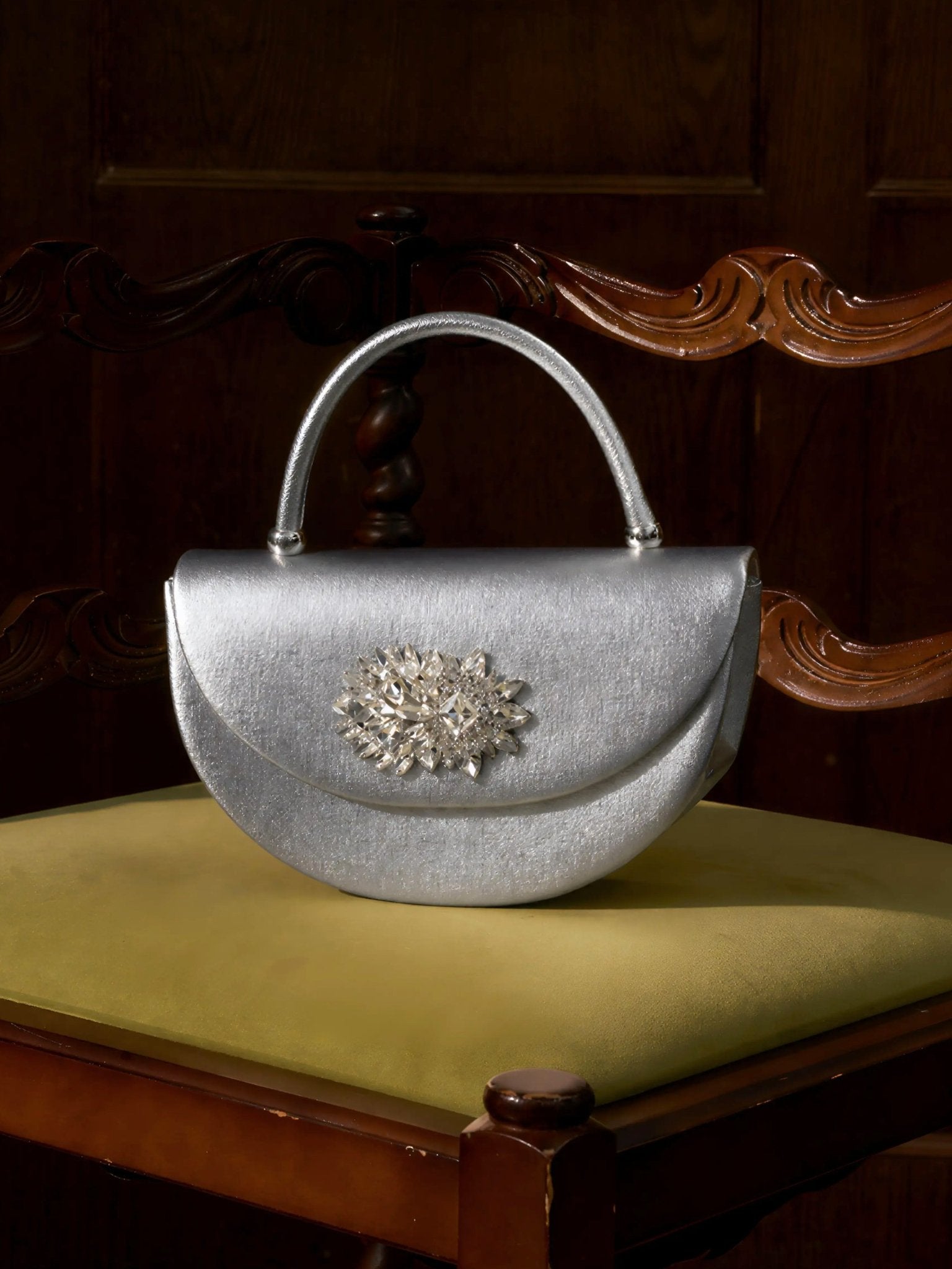 The Social Queen Crystal Floral Satin Mini Top-Handle Bag, featuring a crystal floral embellishment and satin finish, rests on the yellow seat of a carved wooden chair.