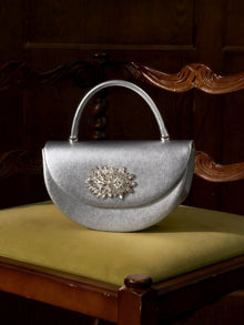 The Social Queen Crystal Floral Satin Mini Top-Handle Bag, featuring a crystal floral embellishment and satin finish, rests on the yellow seat of a carved wooden chair.
