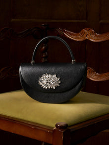 The Social Queen Crystal Floral Satin Mini Top-Handle Bag with a jeweled brooch sits on a yellow-cushioned, carved wooden chair.