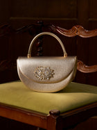 The Social Queen Crystal Floral Satin Mini Top-Handle Bag, featuring jeweled brooch and crystal floral details, rests on an olive green cushioned wooden chair set against a dark wooden background.