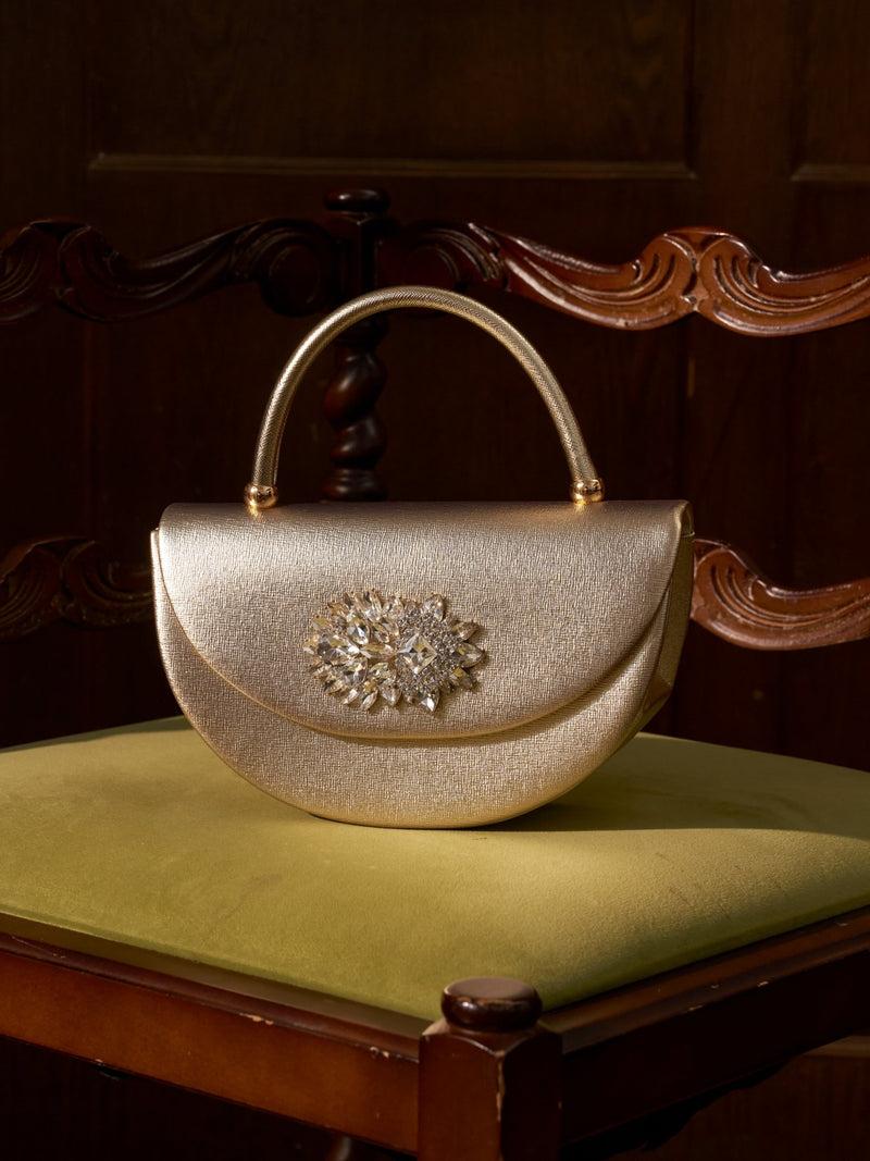 The Social Queen Crystal Floral Satin Mini Top-Handle Bag, featuring jeweled brooch and crystal floral details, rests on an olive green cushioned wooden chair set against a dark wooden background.