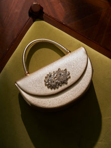 The Social Queen Crystal Floral Satin Mini Top-Handle Bag with a gold textured satin finish and crystal floral embellishment rests on a green velvet chair, bathed in soft light.