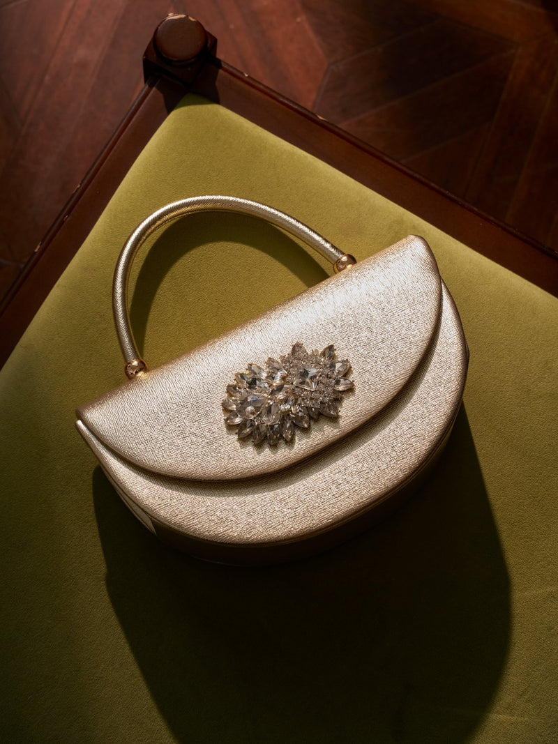 The Social Queen Crystal Floral Satin Mini Top-Handle Bag with a gold textured satin finish and crystal floral embellishment rests on a green velvet chair, bathed in soft light.