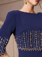 A woman wears the Social Queen Crystal Fringe Fishtail Gown, a navy blue evening dress with gold and silver beaded details and crystal embellishments on the sleeves and bodice; part of her face and an earring are visible.