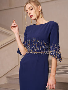 A woman in Social Queen's Crystal Fringe Fishtail Gown stands indoors near a staircase, looking down with one arm raised, the crystal fringe on the sleeves and waist shimmering softly.