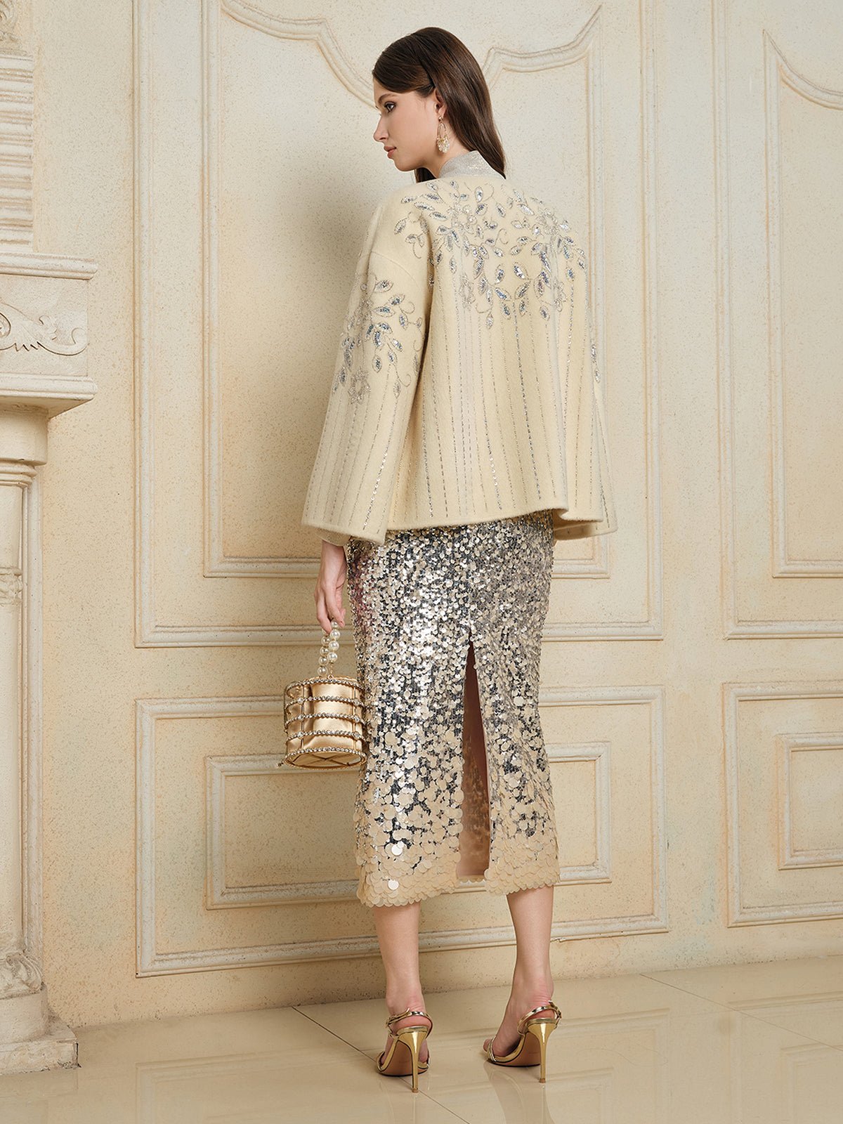 A woman stands indoors facing away, wearing the Social Queen Crystal Leaf Embroidered Wool Coat, a silver sequin leaf-patterned skirt with a slit, gold heels, and holding a gold handbag.