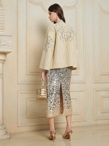 A woman stands indoors facing away, wearing the Social Queen Crystal Leaf Embroidered Wool Coat, a silver sequin leaf-patterned skirt with a slit, gold heels, and holding a gold handbag.