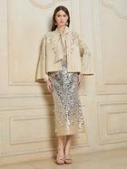 Indoors, a woman wears the Social Queen Crystal Leaf Embroidered Wool Coat over a gold bow-tie blouse, a silver leaf-sequin midi skirt, and gold sandals.