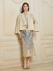 Indoors, a woman wears the Social Queen Crystal Leaf Embroidered Wool Coat over a gold bow-tie blouse, a silver leaf-sequin midi skirt, and gold sandals.