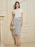 Woman poses indoors against an ornate cream wall in Social Queen's Crystal & Sequin Knit Textured Crop Top, paired with a silver fringed skirt, gold sandals, and a beige purse.