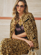 A woman seated, wearing sunglasses, a black top, gold jewelry, and the Social Queen Crystal-Trim Sequin Velvet Jacket with matching pants, holding a small gold handbag in her lap.