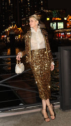 By the waterfront at night, a person wears Social Queen’s Sequin Velvet Skirt with a matching jacket and sheer white blouse, holding a sparkly handbag. City lights and buildings shimmer in the background.